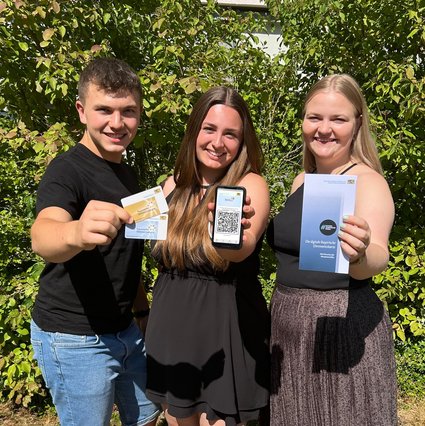Drei junge Personen halten blaue und goldene Ehrenamtskarte, ein Smartphone und einen Flyer zur Ehrenamtskarte in die Kamera