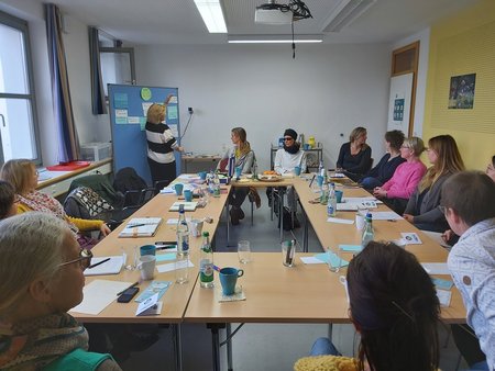 Gruppe von Personen sitzt um Tisch, eine Person steht am Flipchart
