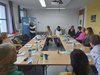 Gruppe von Personen sitzt um Tisch, eine Person steht am Flipchart